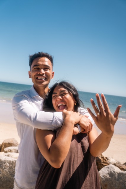 Couple celebrating their engagement, showing the ring in a joyful embrace at the beach